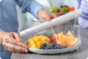 Plastic wrap covering a disposable fruit tray with sliced oranges, mango, and blueberries.